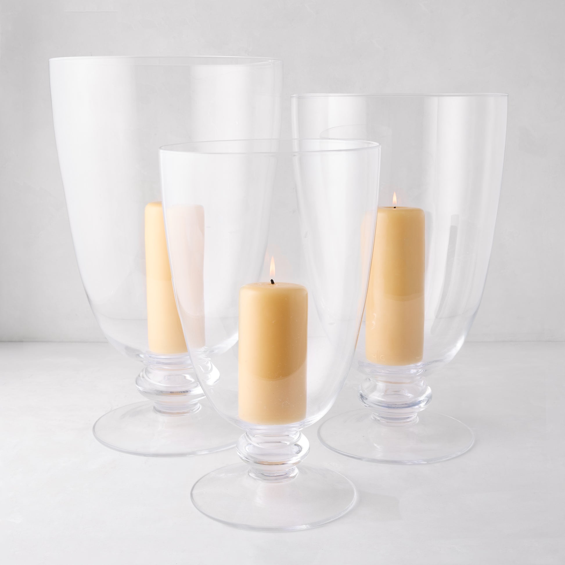 Three clear Mayfair glass hurricanes of varying sizes with lit cream candles inside, on a white surface.