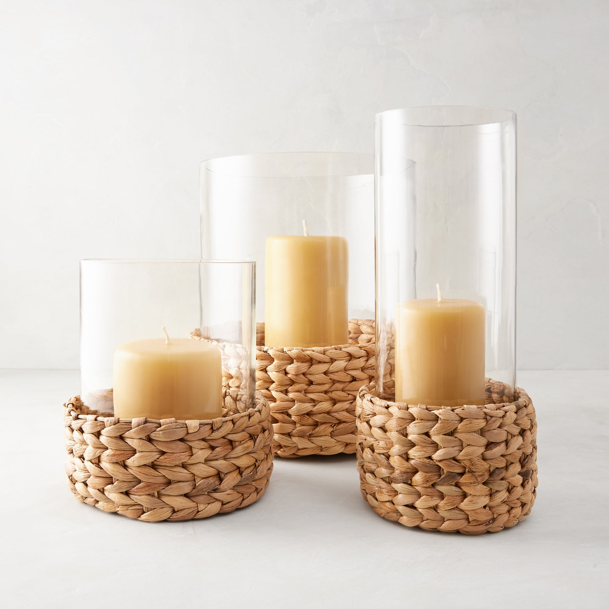 Three glass hurricanes with woven natural hyacinth bases and amber candles.