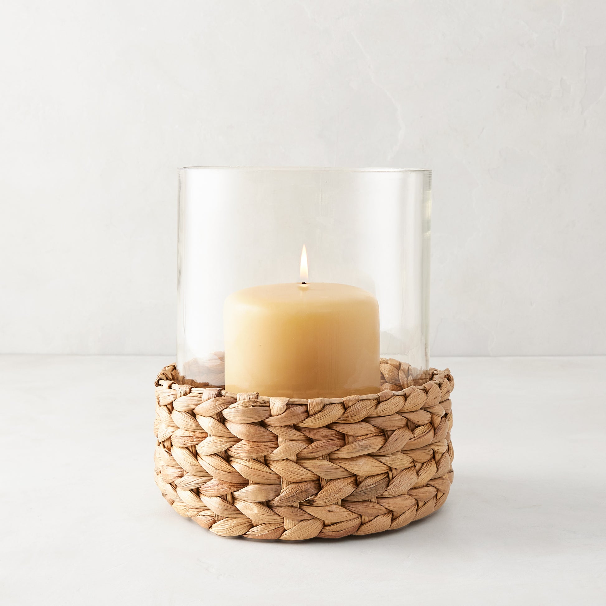 Medium glass hurricane with a woven natural hyacinth base and lit amber candle.