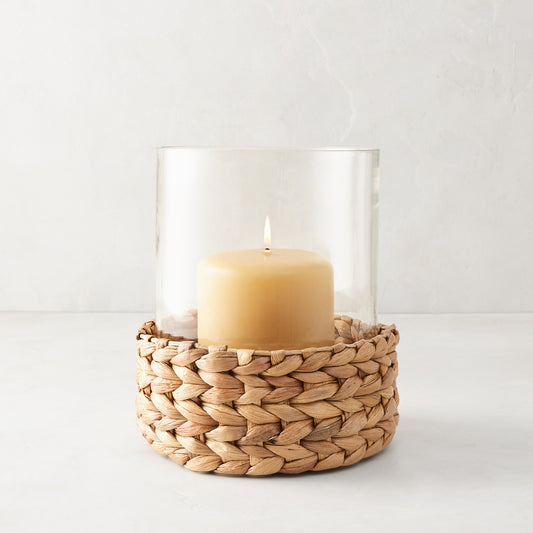 Medium glass hurricane with a woven natural hyacinth base and lit amber candle.