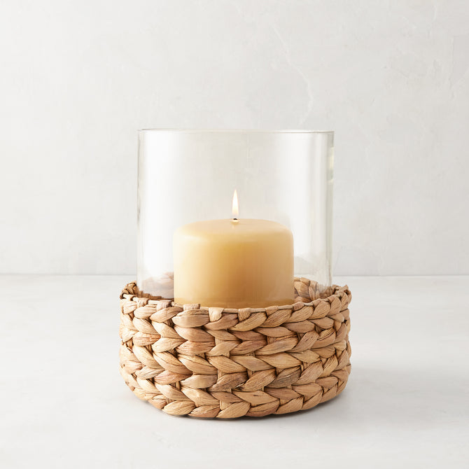 Medium glass hurricane with a woven natural hyacinth base and lit amber candle.