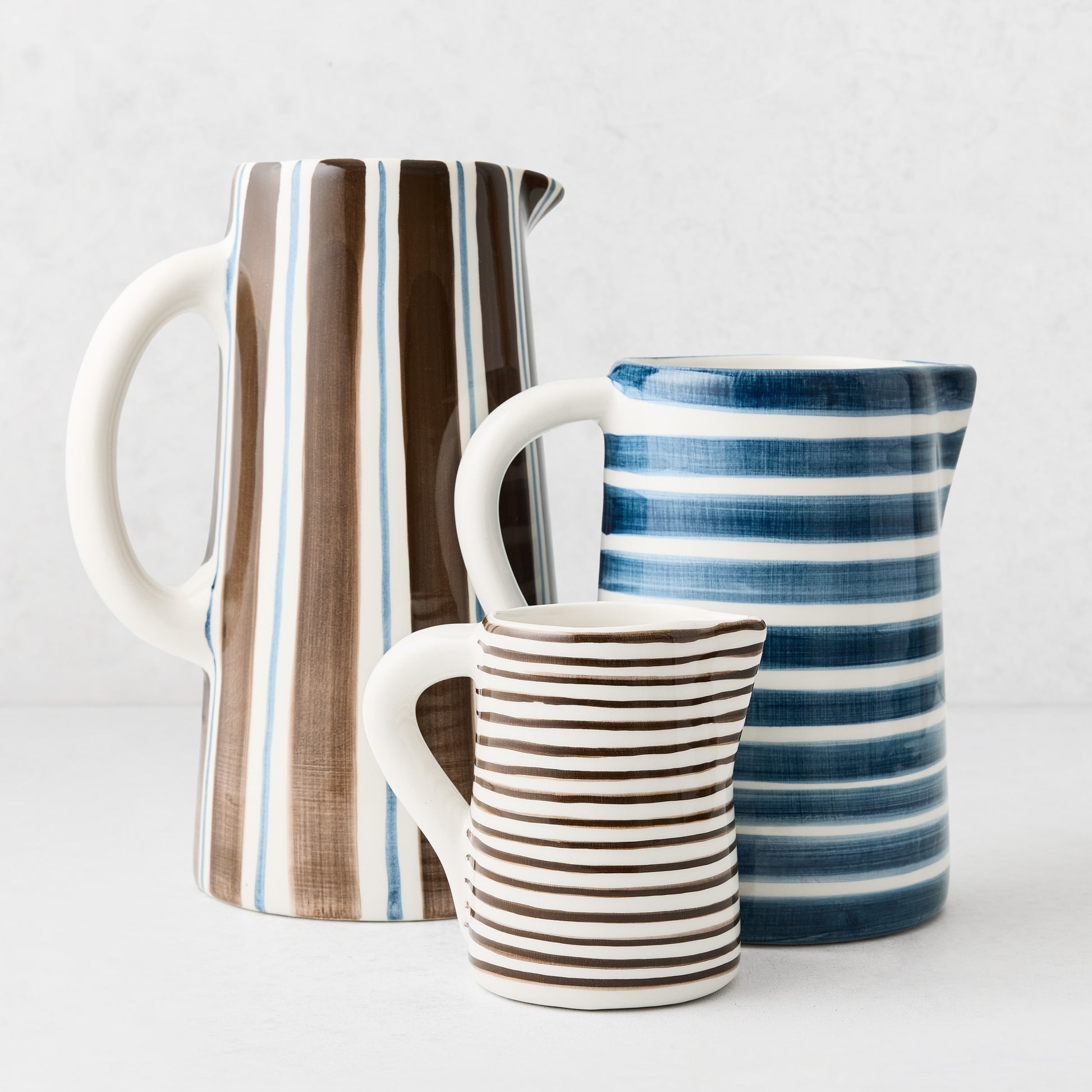Three ceramic pitchers in varying sizes with hand-painted blue and brown striped patterns on a white background.
