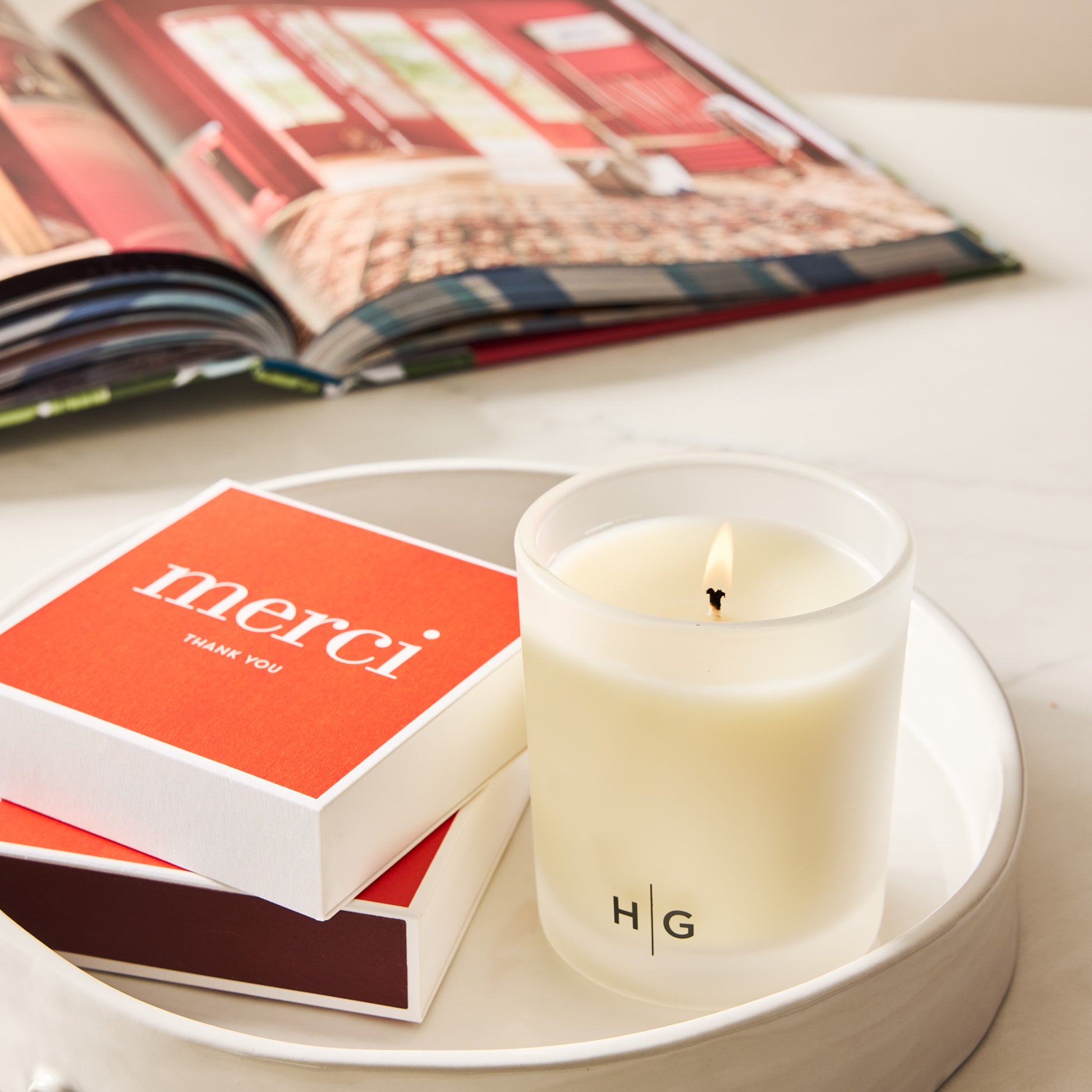 Red "merci" boxed matches with white lettering next to a lit Hudson Grace candle on a white tray.
