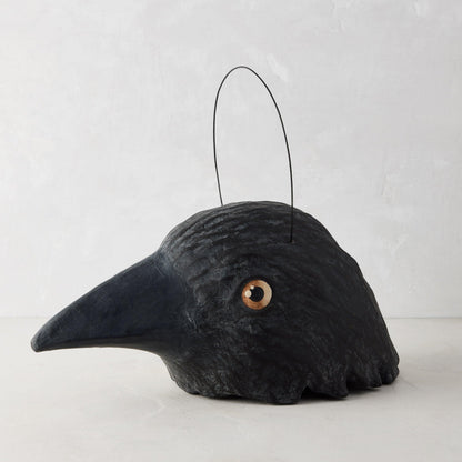 Black papier-mâché raven candy bucket with a distressed finish, wire handle, and vintage-style eye, 17 inches long.