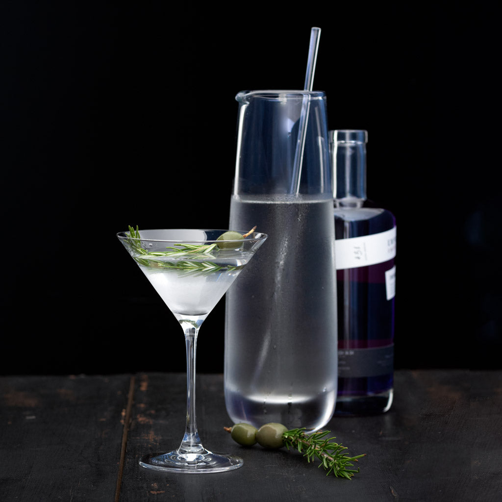 Clear Midtown Martini Glass with a drink, rosemary, and olives, next to a glass carafe and purple bottle.