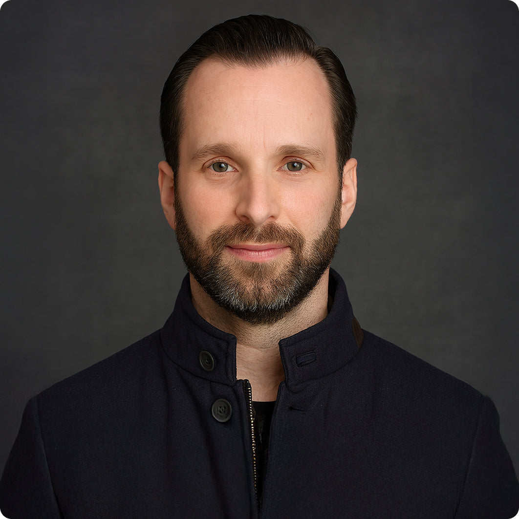 Bearded man with dark hair wearing a navy blue high-collar jacket against a dark gray background.