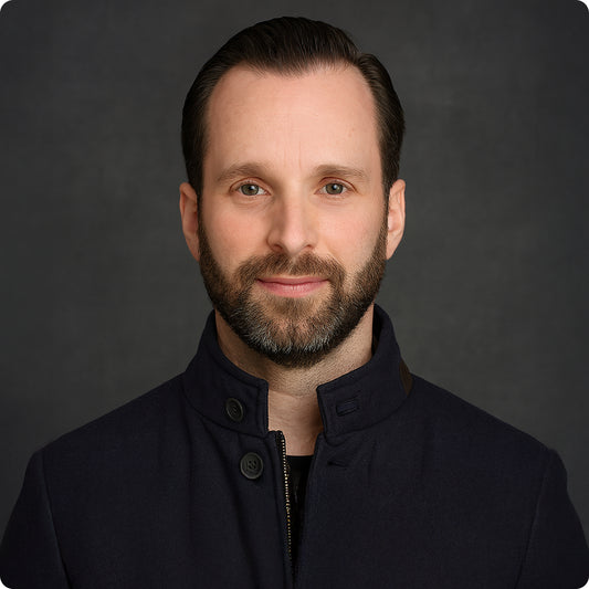 Bearded man with dark hair wearing a navy blue high-collar jacket against a dark gray background.