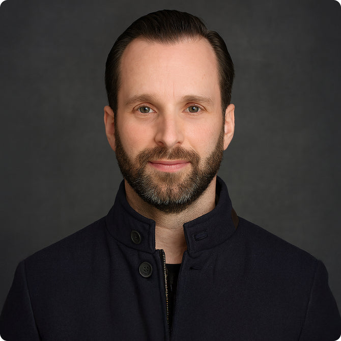 Bearded man with dark hair wearing a navy blue high-collar jacket against a dark gray background.