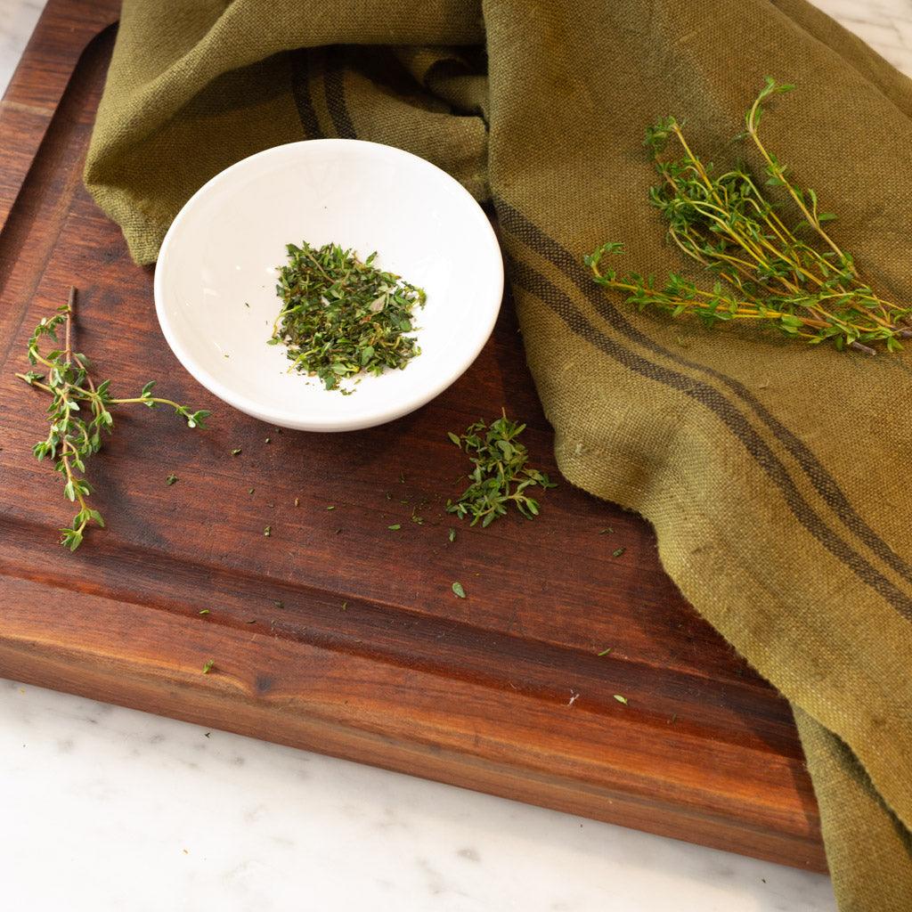 White ceramic dipping bowl with dried herbs on a dark wood cutting board and green linen.