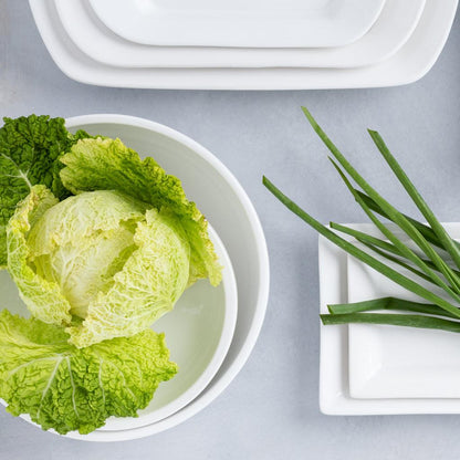 White ceramic serve bowl with green cabbage, next to white plates and green onions.