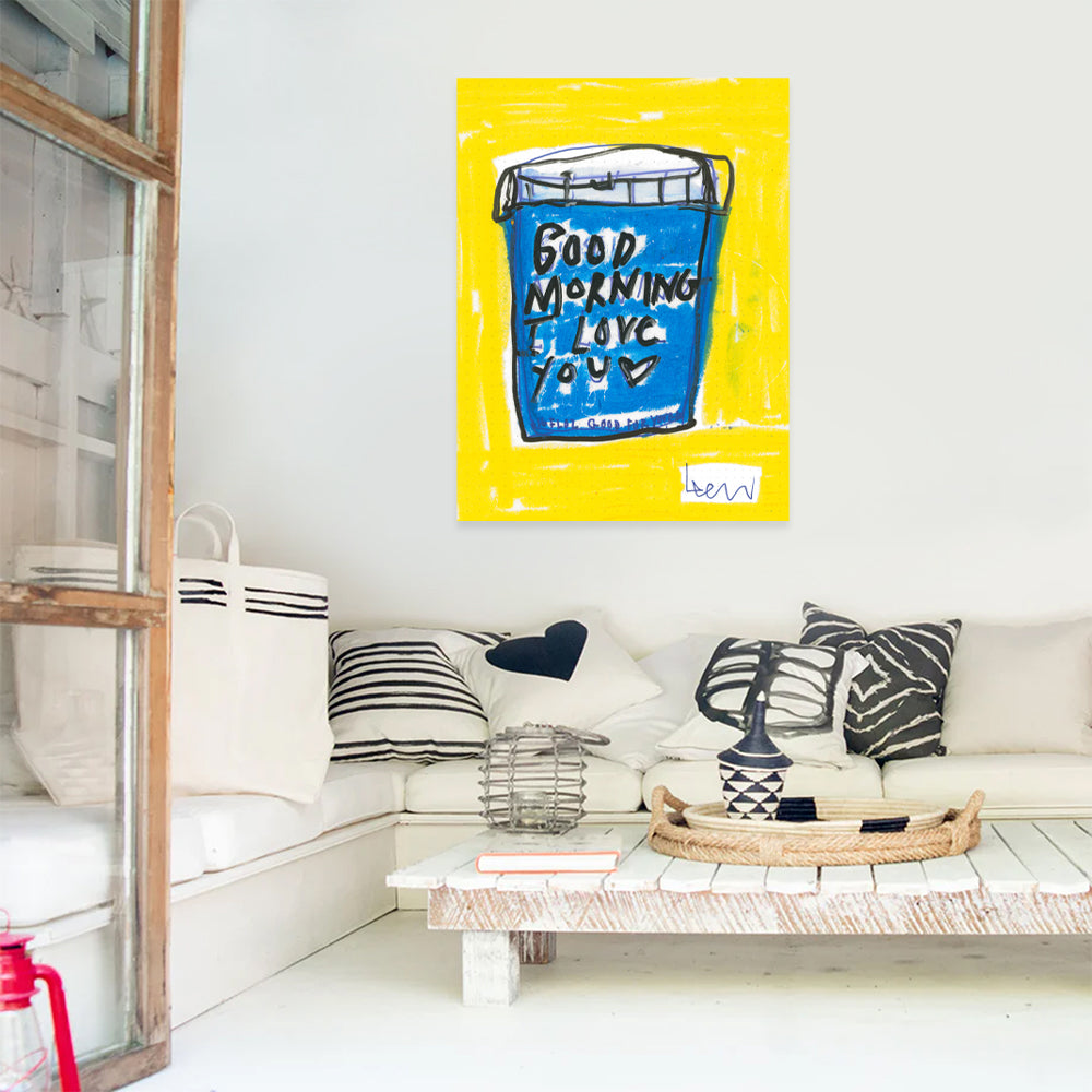 Kerri Rosenthal's "My First Love" art print, a vibrant blue cup on a yellow background, framed and hung above a white sofa.
