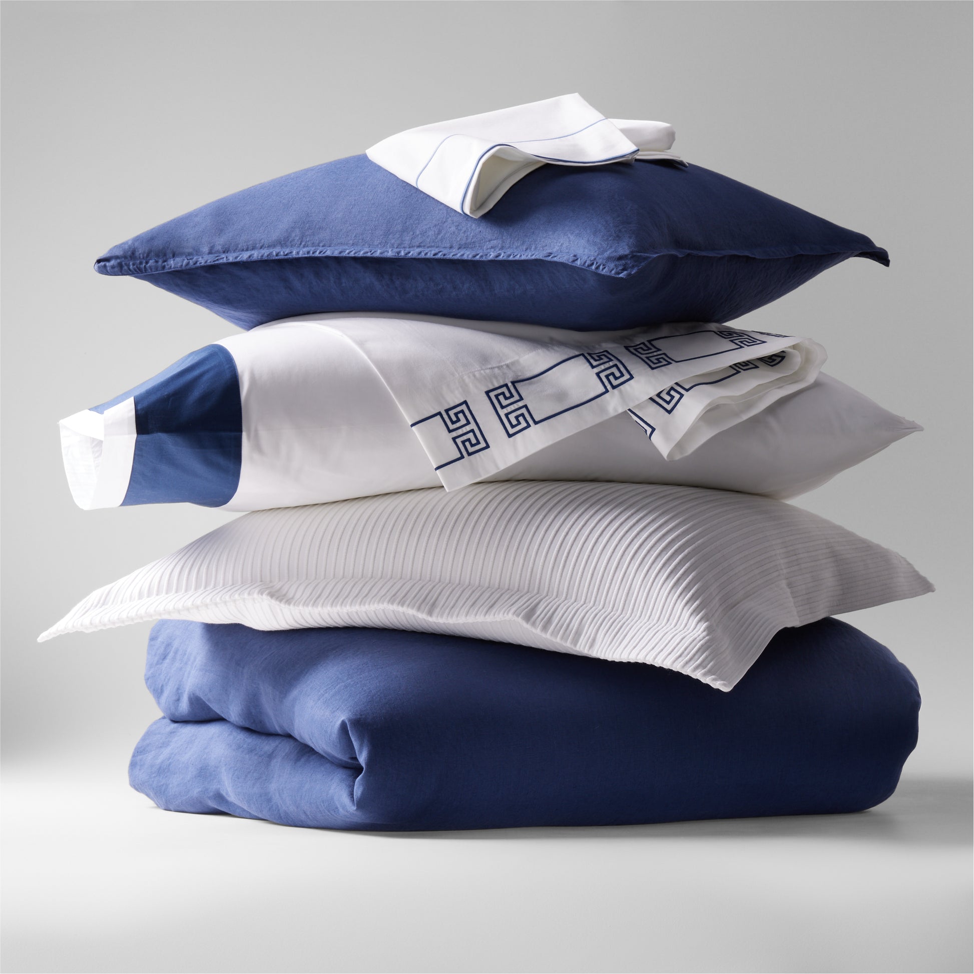 Stack of navy blue washed linen duvet cover, white pleated pillow, and white percale sheets with blue trim.