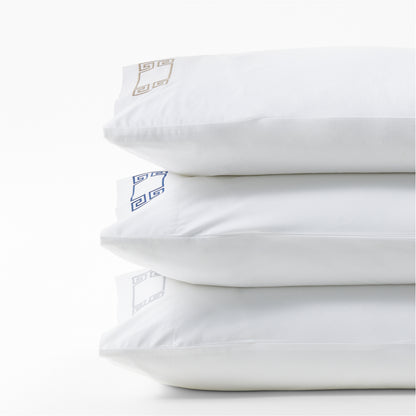 Stack of three white percale pillowcases with embroidered Greek key designs in navy, gold, and silver.