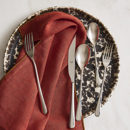 Neve 5-piece mirrored stainless steel flatware set with slim handles on a patterned plate and rust-colored linen napkin.