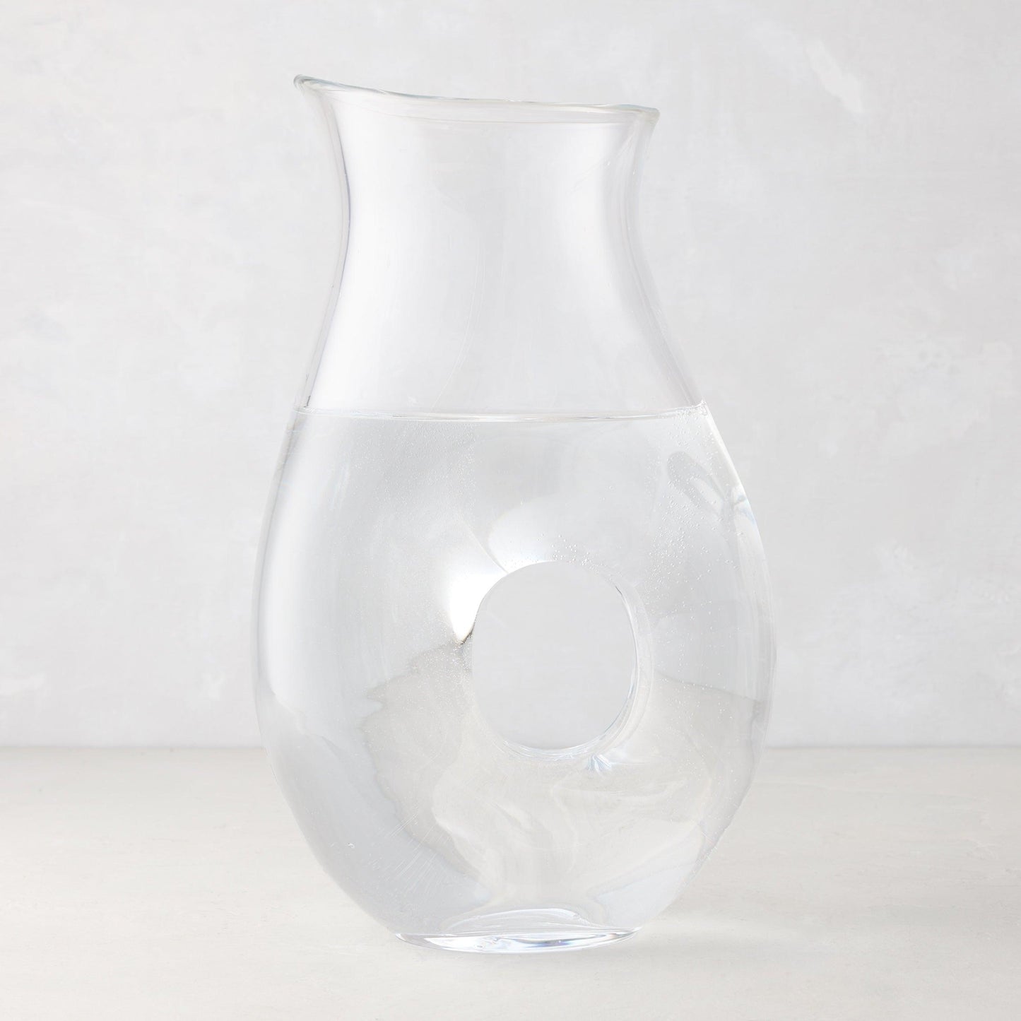 Clear glass "O" pitcher with an O-shaped handle, filled with water, on a light background.