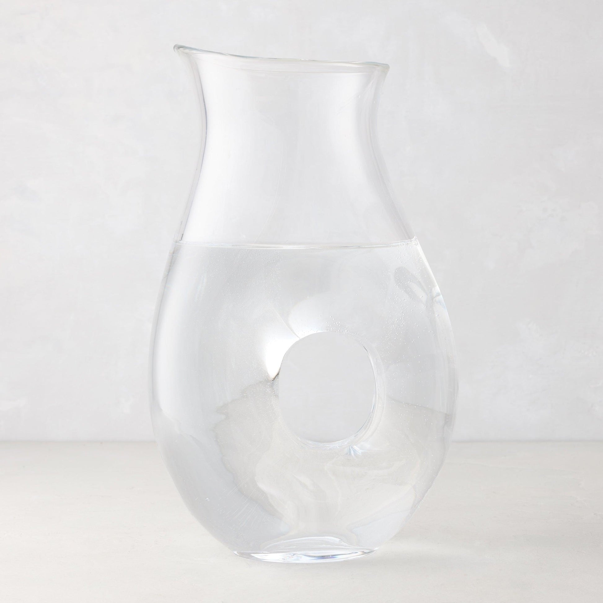 Clear glass "O" pitcher with an O-shaped handle, filled with water, on a light background.