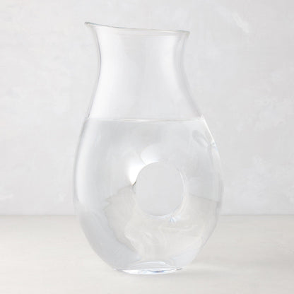 Clear glass "O" pitcher with an O-shaped handle, filled with water, on a light background.