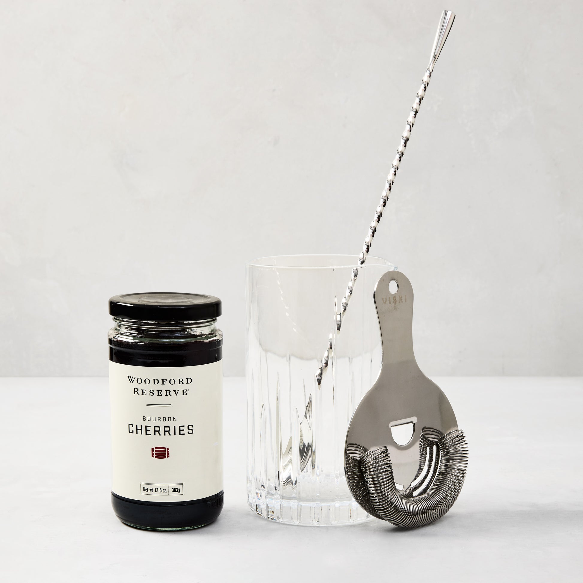 Woodford Reserve Bourbon Cherries jar, clear ribbed glass, silver cocktail stirrer, and stainless steel strainer.