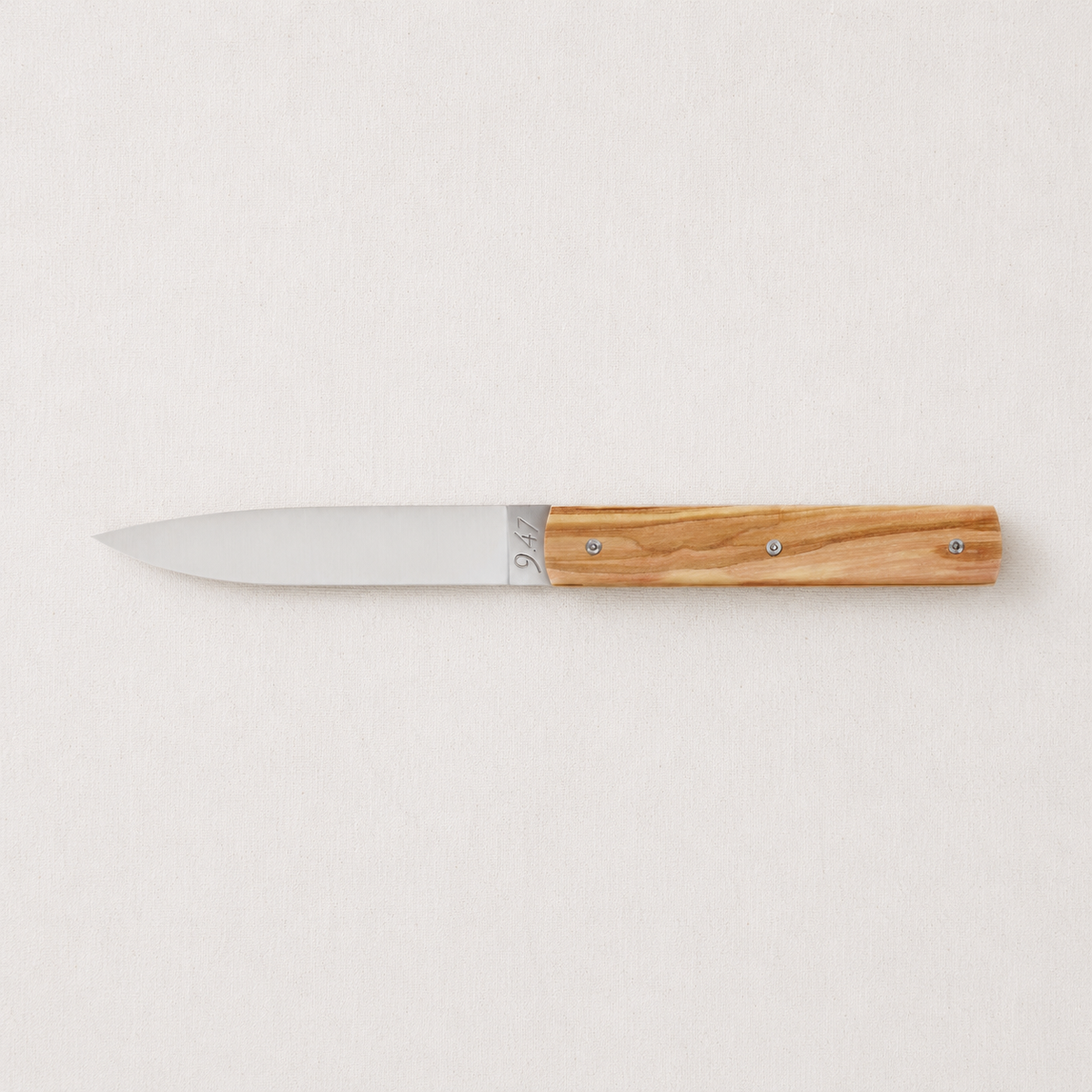 Perceval 9.47 stainless steel knife with a natural olive wood handle on a light textured background.