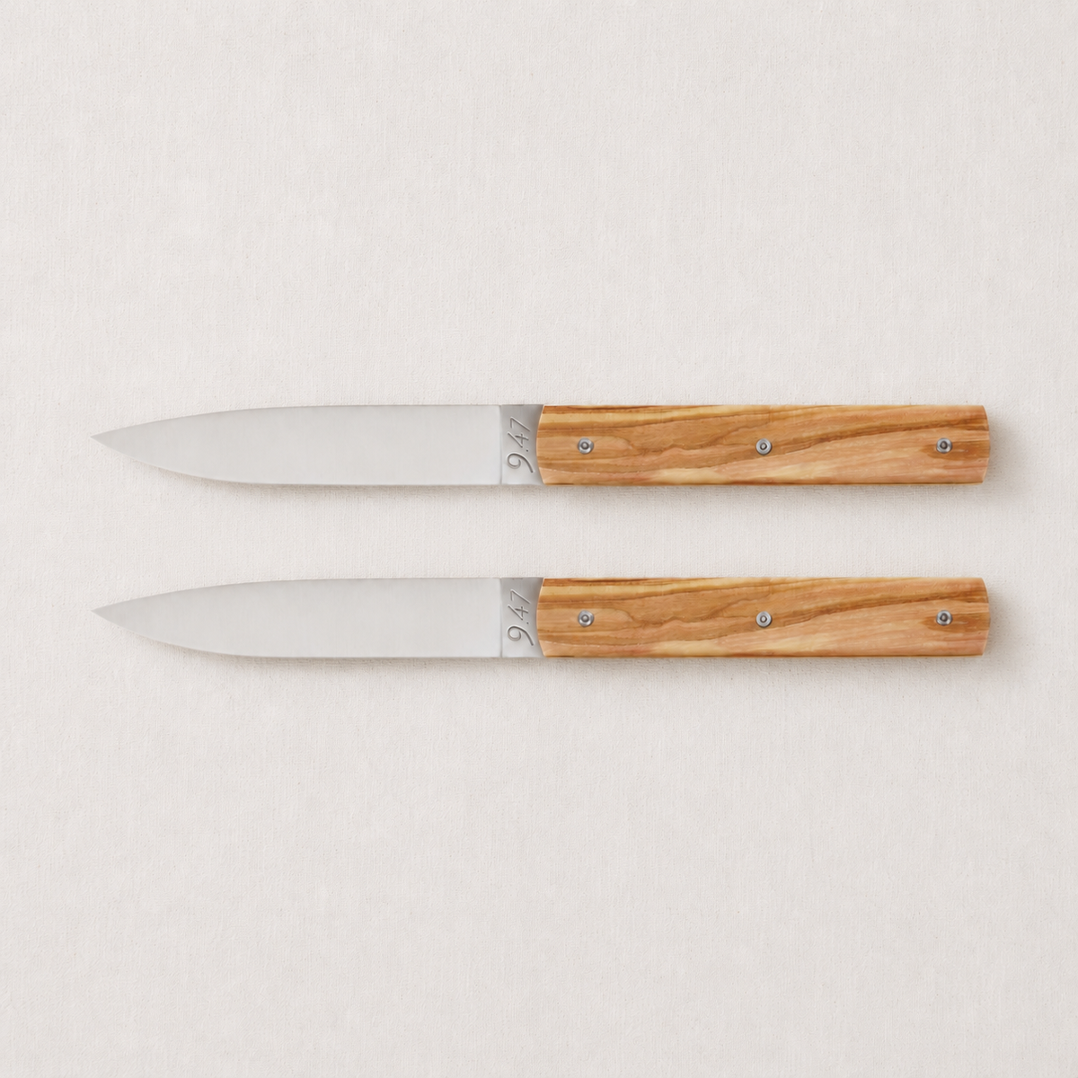 Set of two Perceval 9.47 knives with stainless steel blades and polished olive wood handles on a light background.