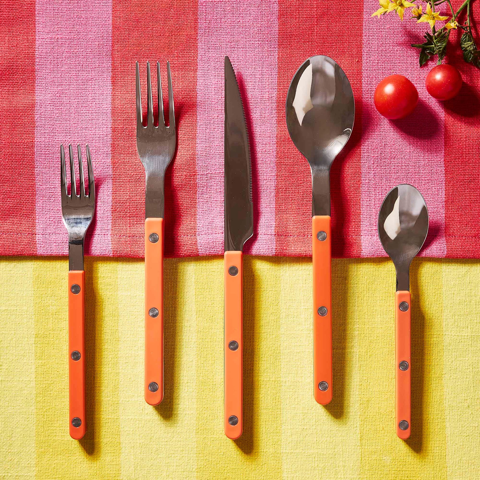 Orange 5-piece bistro flatware set with stainless steel heads and riveted acrylic handles on a striped tablecloth.
