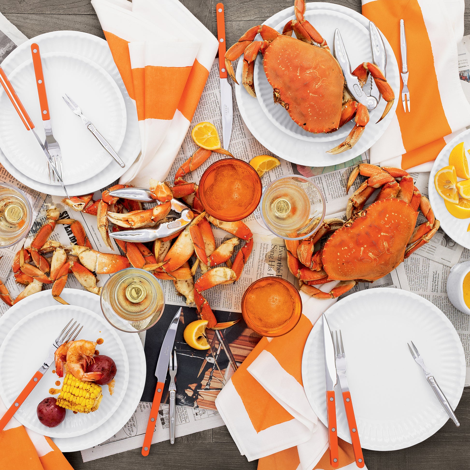 Overhead view of a crab boil with orange-handled Bistro flatware, white plates, drinks, and newspaper.