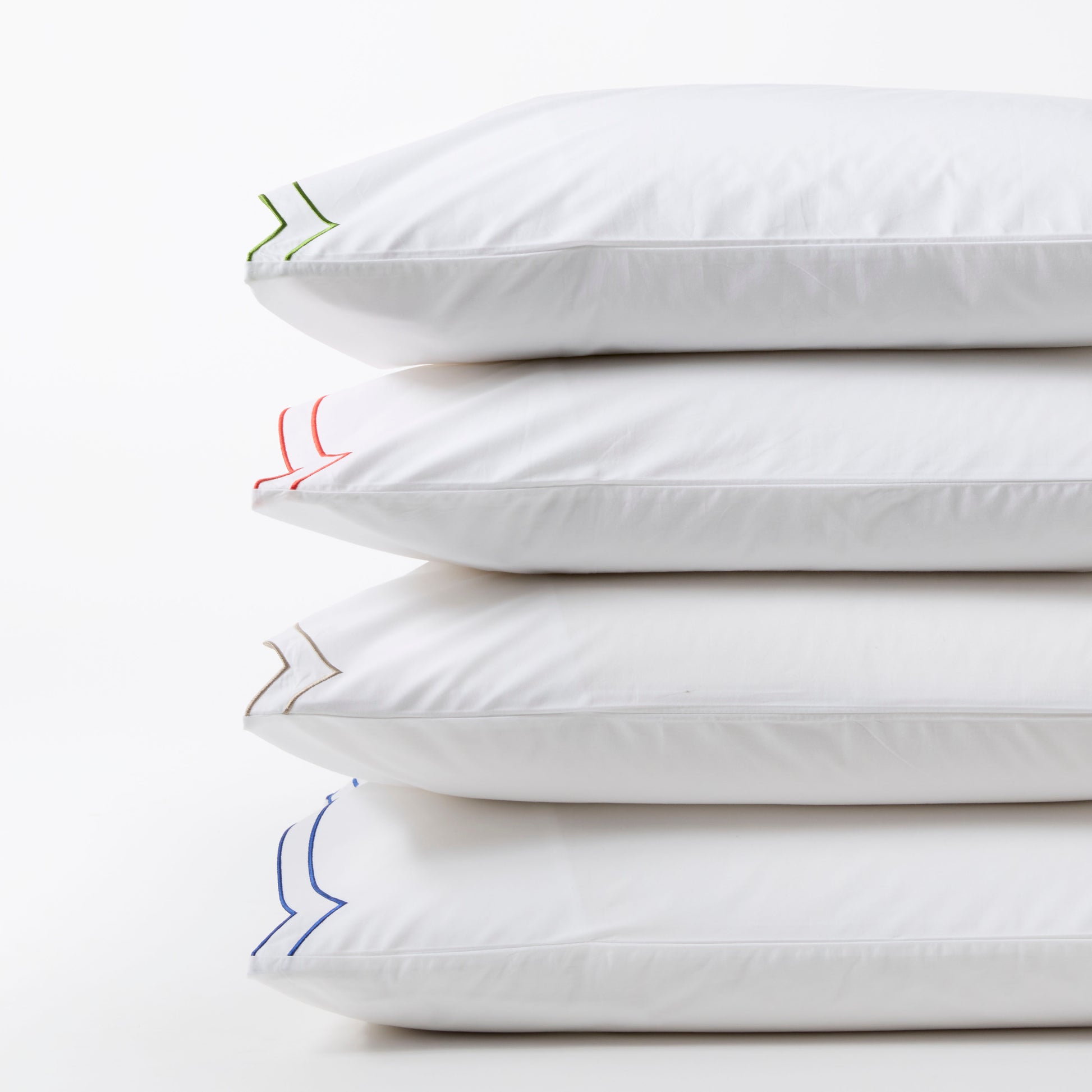 Stack of four white organic cotton percale pillow shams with double scallop stitch embroidery in green, orange, tan, and blue.