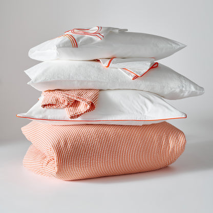 Stack of white percale Euro shams with orange piping, folded orange striped duvet, and matching bedding.