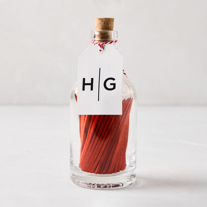 Glass bottle with cork stopper, filled with red matches, and a white HG tag tied with red and white string.