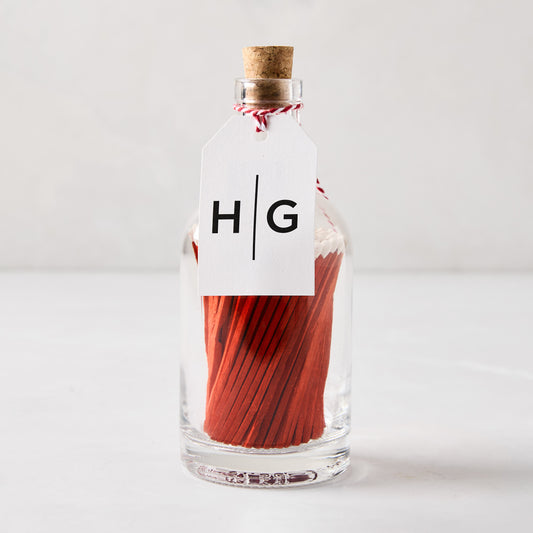 Glass bottle with cork stopper, filled with red matches, and a white HG tag tied with red and white string.