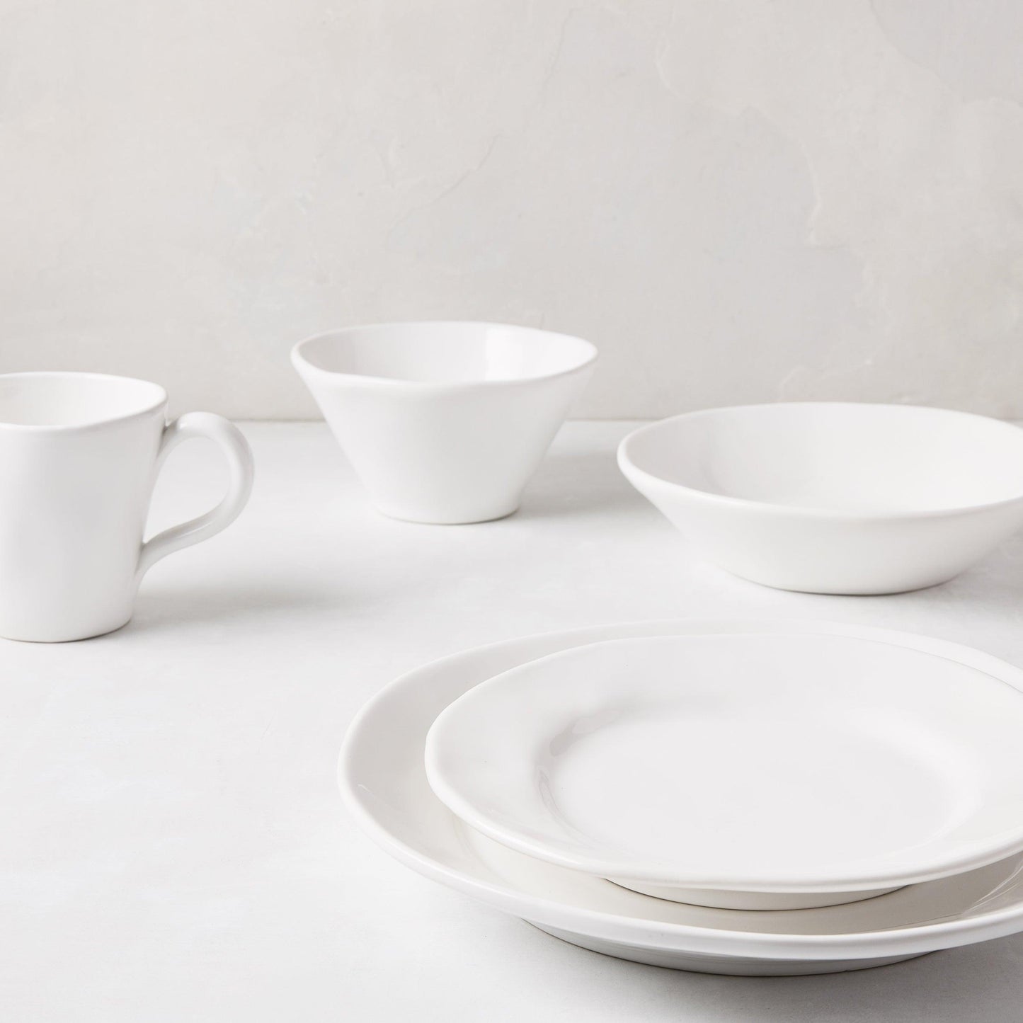 Stack of two creamy white ceramic dinner plates with subtle organic edges, a bowl, and a mug.