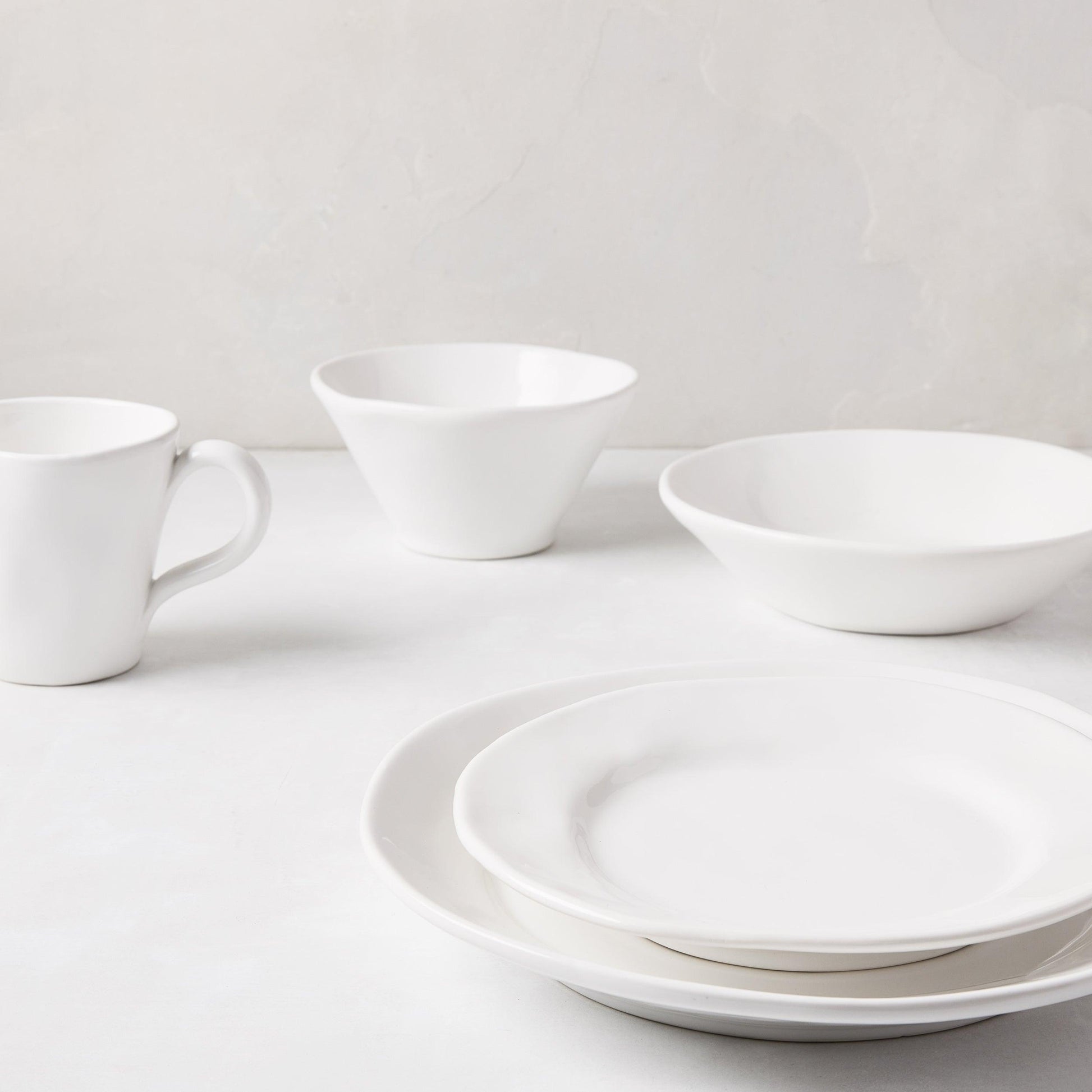 Stack of two creamy white ceramic dinner plates with subtle organic edges, a bowl, and a mug.