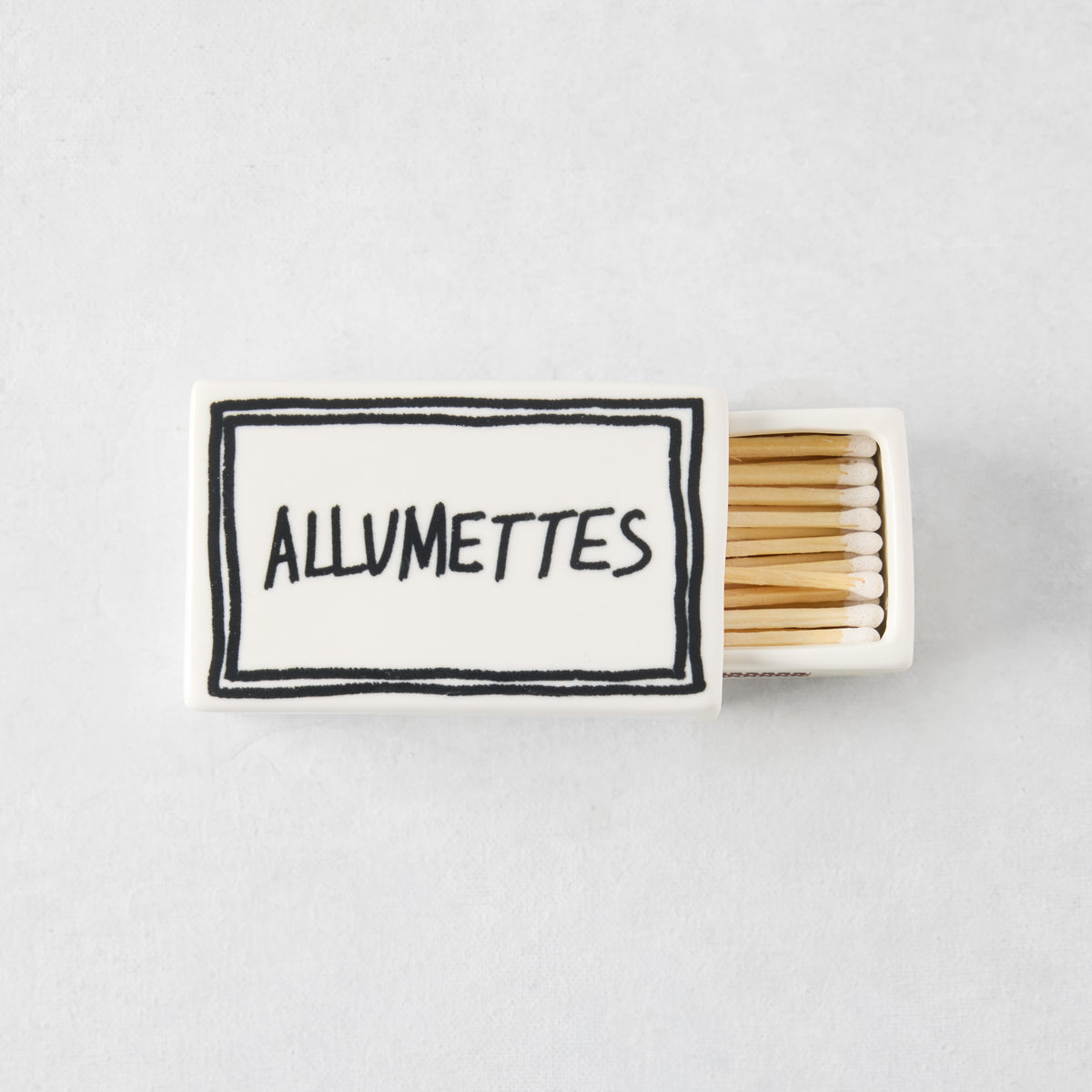 White ceramic match box with hand-drawn black border and Allumettes text, filled with wooden matches.