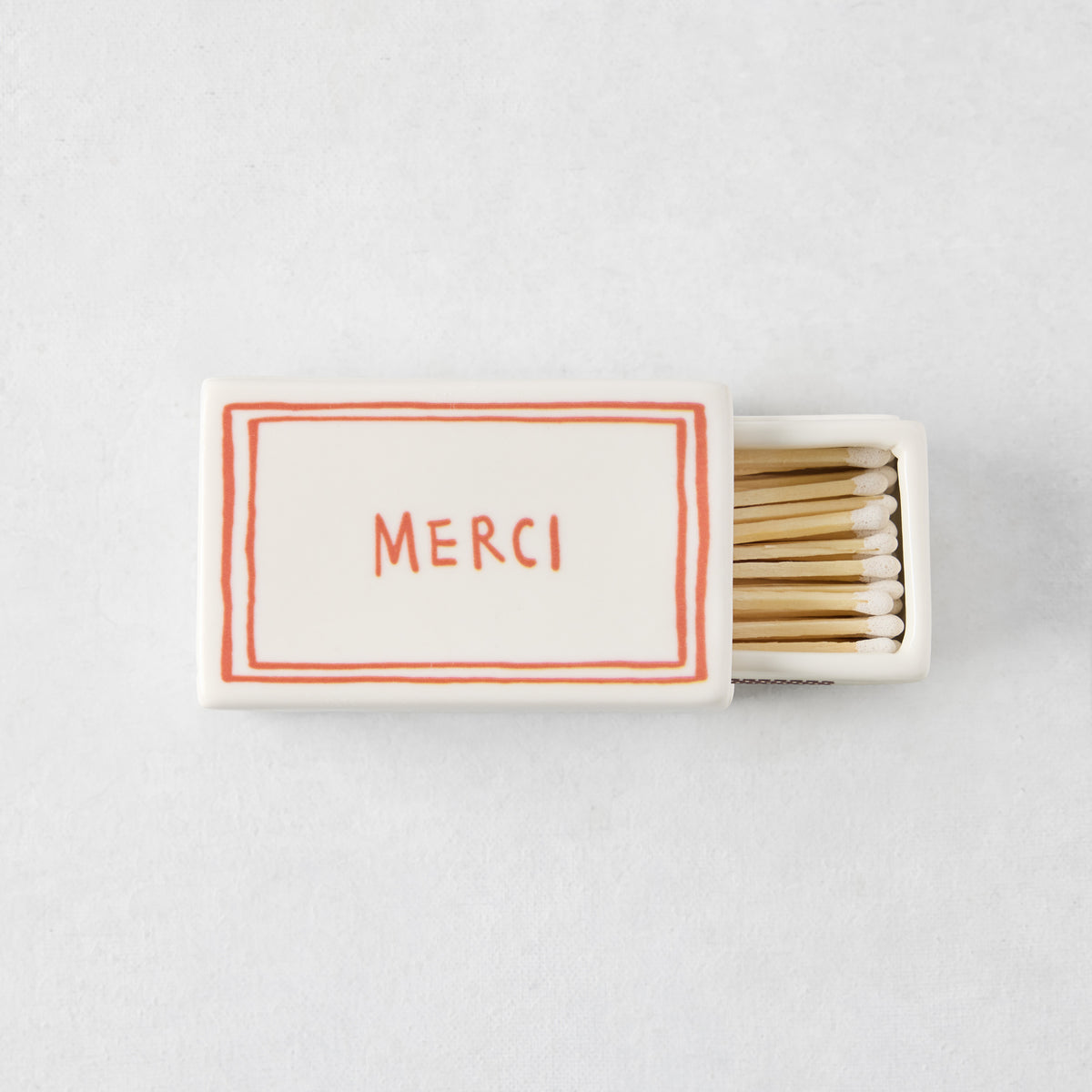 White porcelain match box with red border and Merci text, filled with wooden matches, 3.54 by 2.17 inches.