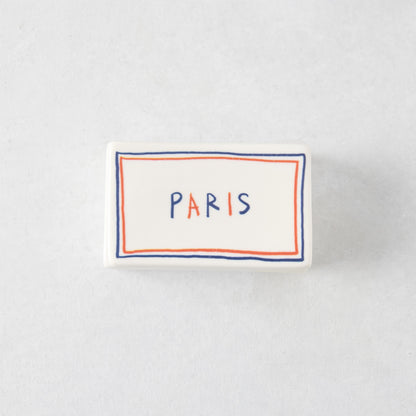 Rectangular white ceramic match box with a hand-painted blue and orange border and the word PARIS in colorful letters.