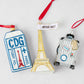 Parisian-themed Christmas ornament set featuring a glittery CDG luggage tag, gold Eiffel Tower, and silver suitcase.