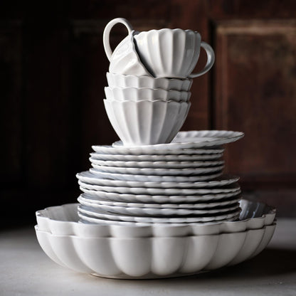White glazed stoneware mug with a scalloped petal design, stacked atop matching ceramic plates and bowls.