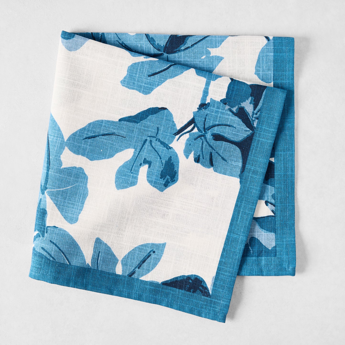 Peter Dunham + Hudson Grace Blue Fig Leaf printed white cotton slub napkin with blue border, folded.