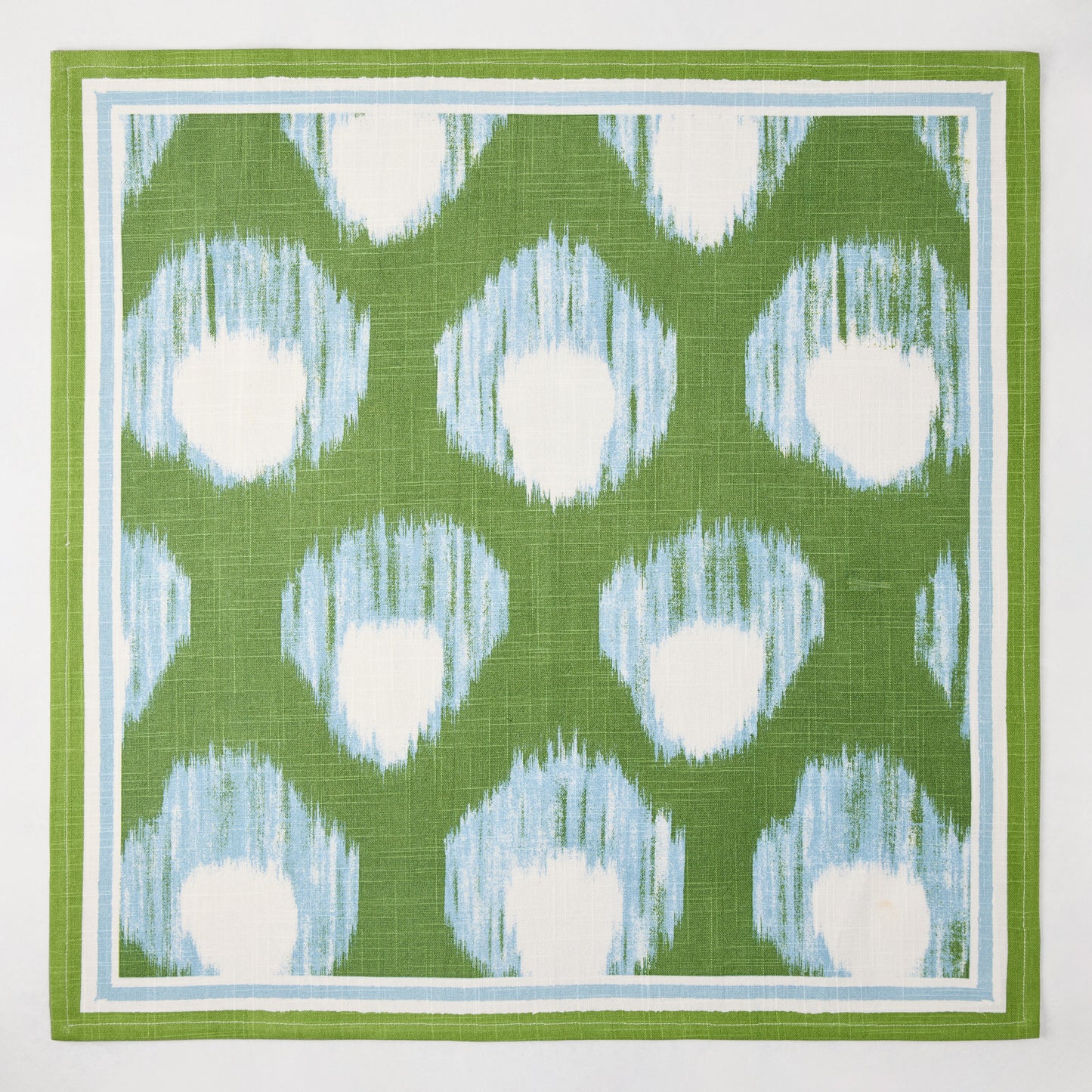 Green Bukhara Ikat printed cotton slub napkin with blue and white blurred pattern and border.