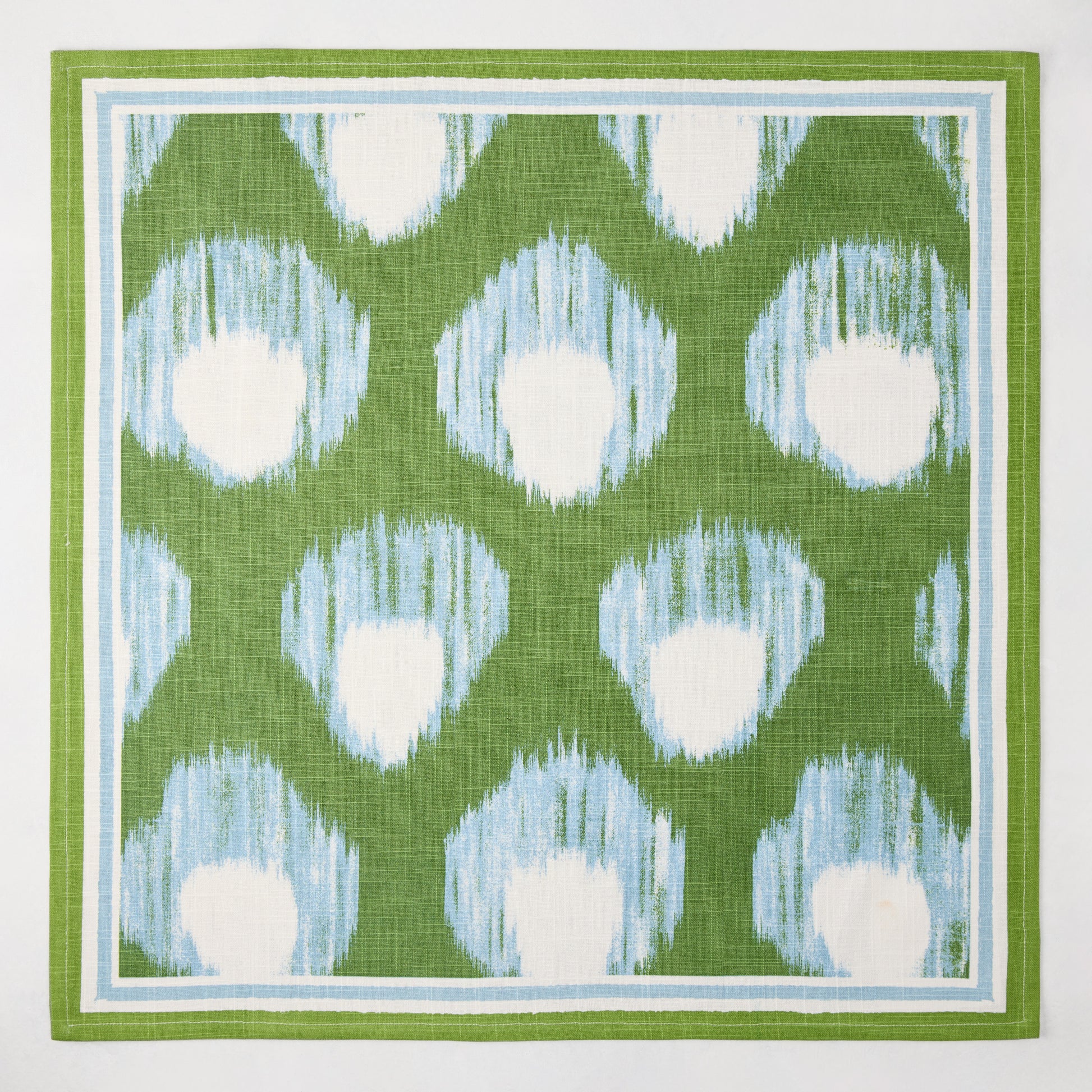 Green Bukhara Ikat printed cotton slub napkin with blue and white blurred pattern and border.