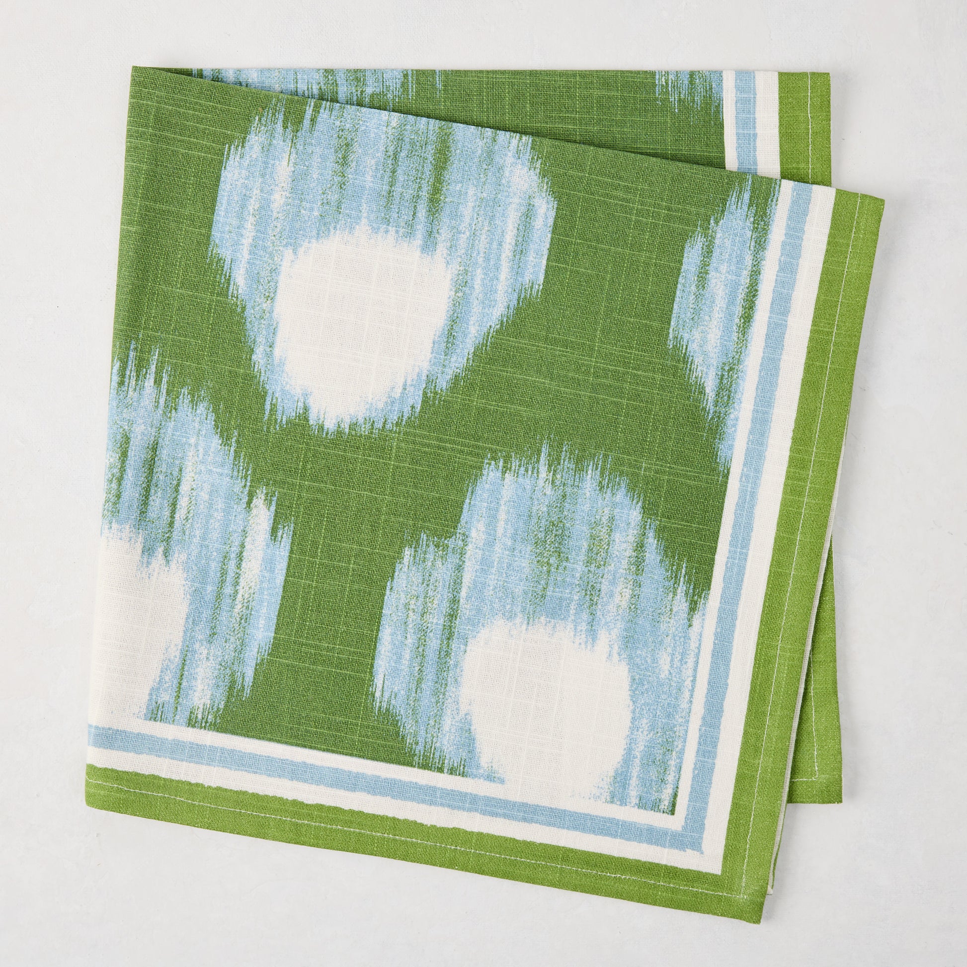 Green Bukhara Ikat Printed Napkin, 100% cotton slub, with blue and white blurred pattern and striped border.
