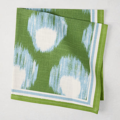 Green Bukhara Ikat printed cotton slub napkin with blue and white blurred pattern and striped border.
