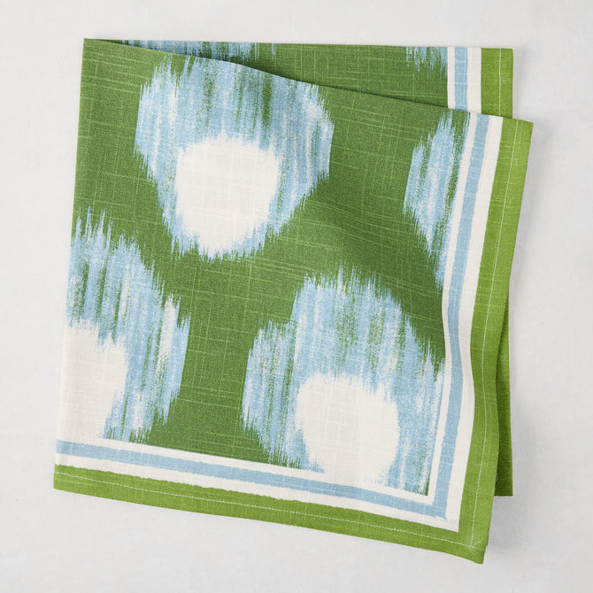 Green Bukhara Ikat printed cotton slub napkin with blue and white blurred pattern and striped border.
