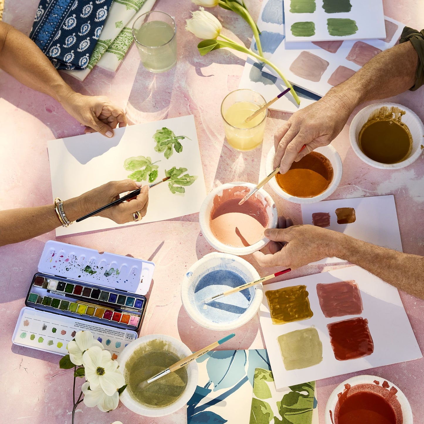 Hands painting green fig leaf designs on paper with watercolors, brushes, and paint palettes on a pink table.