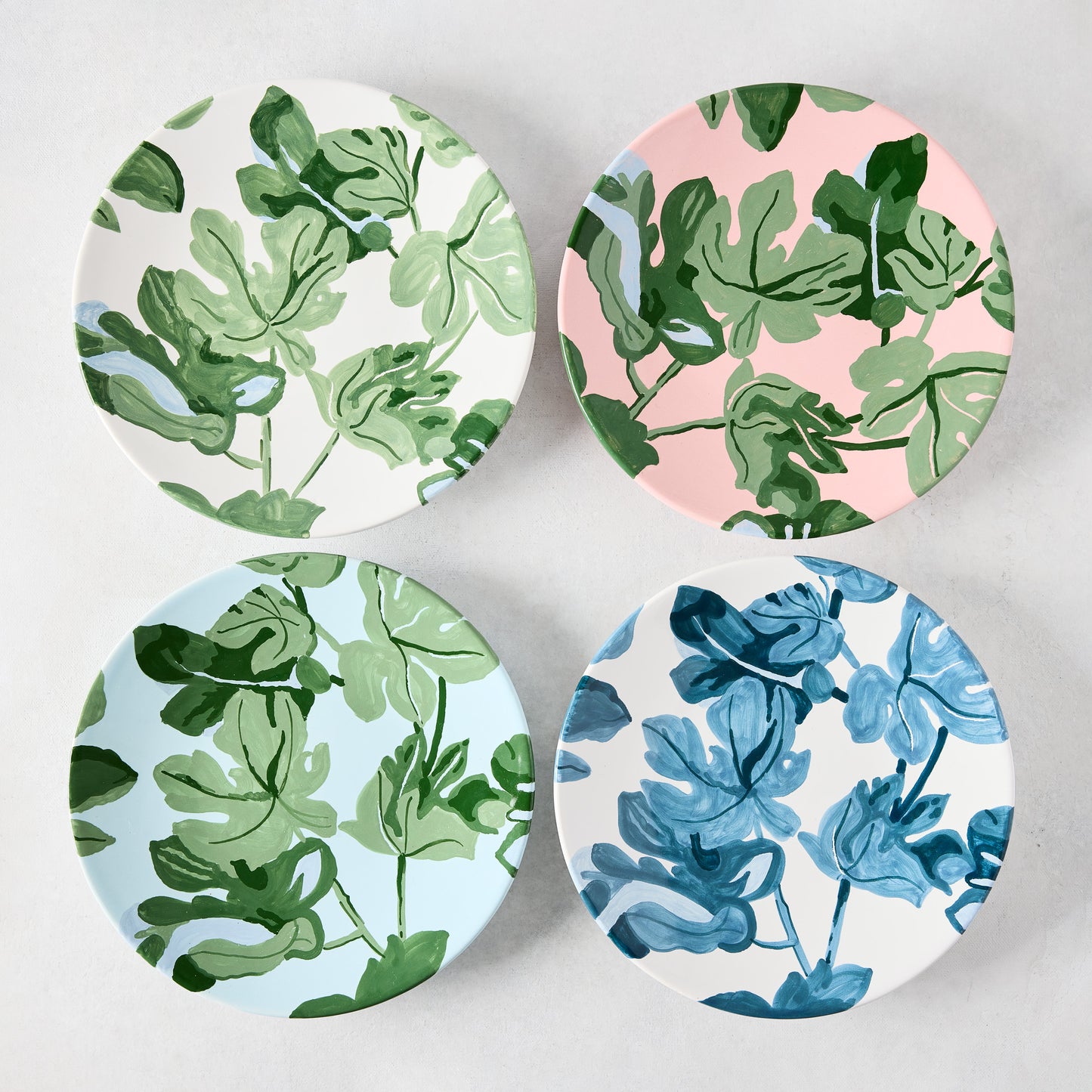 Four ceramic salad plates with hand-painted green and blue fig leaf patterns on white, pink, and light blue backgrounds.