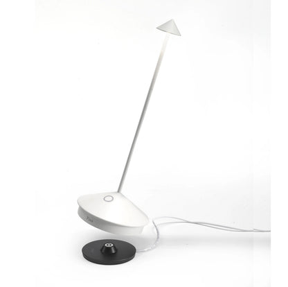 White Pina Pro rechargeable LED lamp with die-cast aluminum base, thin rod, and polycarbonate shade, next to a black charging base.