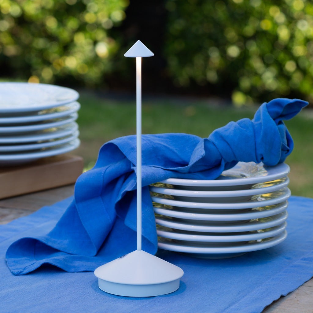 White die-cast aluminum Pina Pro rechargeable LED lamp on a blue placemat, next to stacked white plates.
