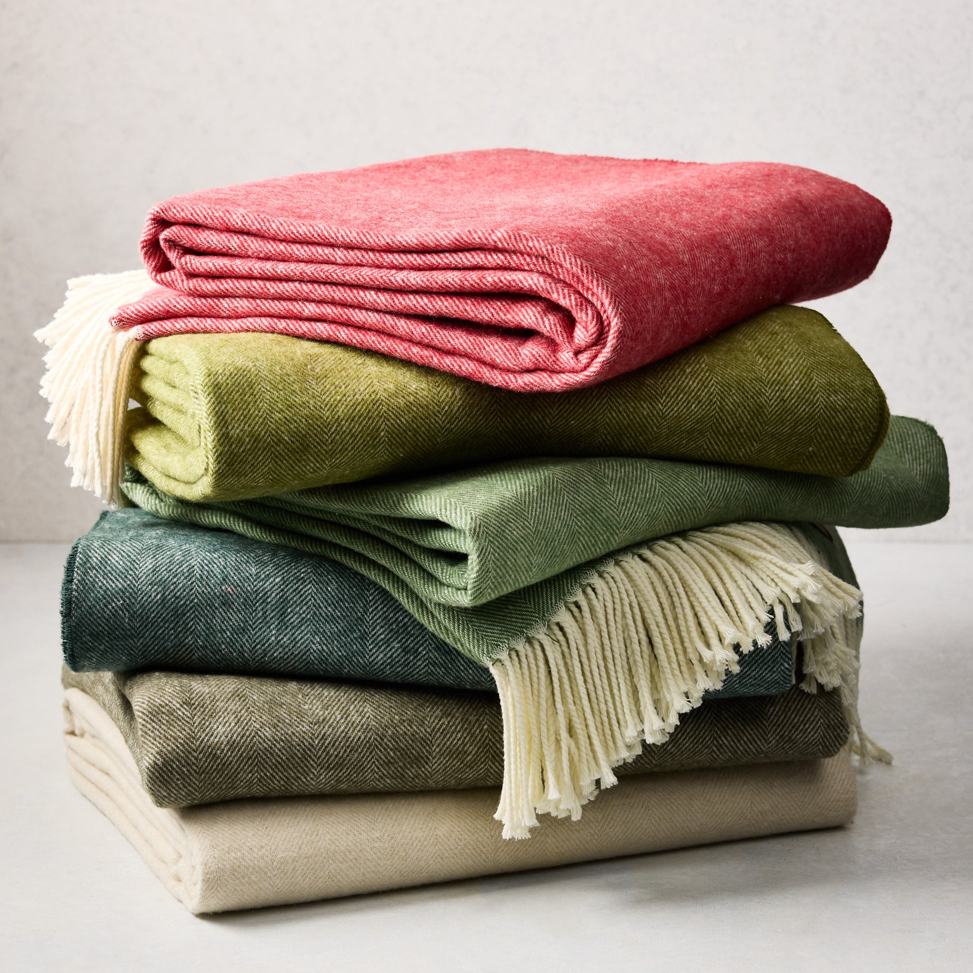 Stack of folded Italian herringbone throw blankets in red, green, blue, olive, and cream with fringe.