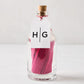 Glass bottle with cork stopper, filled with pink matches, and a white HG tag.