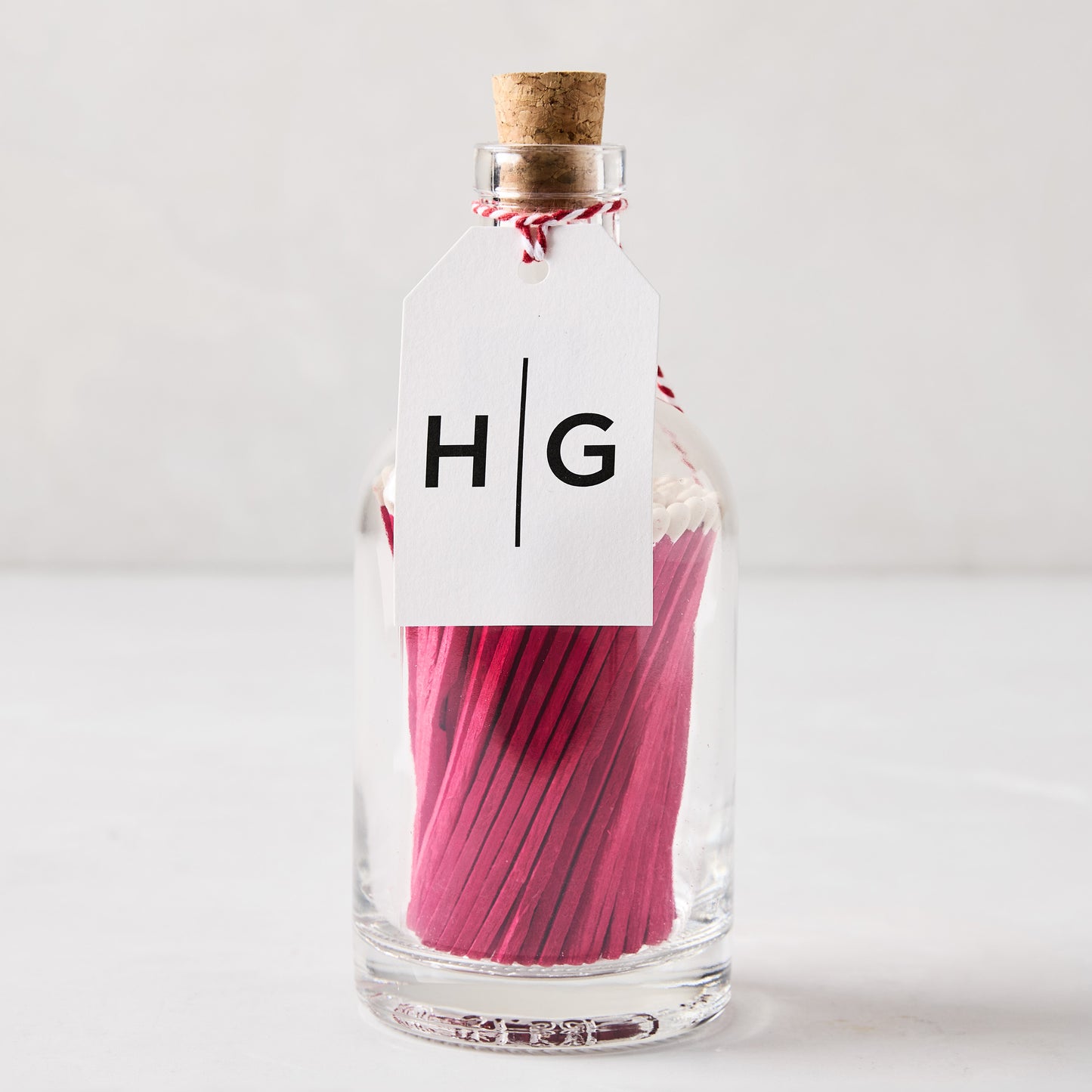 Glass bottle with cork stopper, filled with pink matches, and a white HG tag.